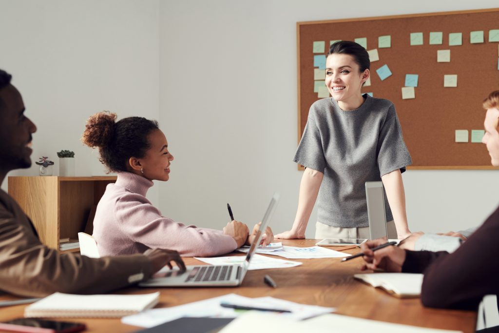 A person smiling and standing at the head of a table, leading a business meeting.